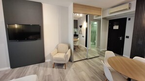 Une chambre moderne dotée d’un téléviseur à écran plat fixé au mur, d’un fauteuil beige, d’une table à manger en bois avec des chaises, et d’une cabine de douche en verre.
