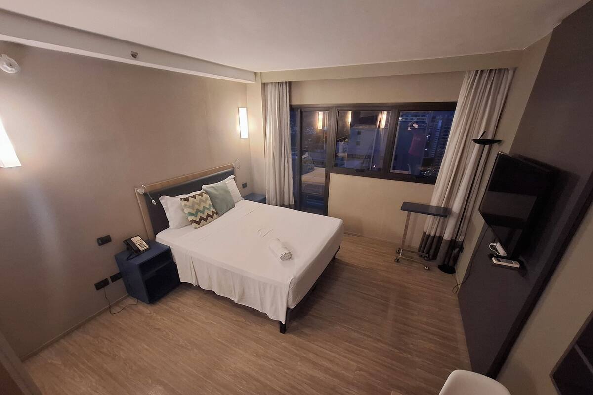 Une chambre d’hôtel comprenant un lit, une table de chevet, une lampe, une fenêtre avec des rideaux et un téléviseur.