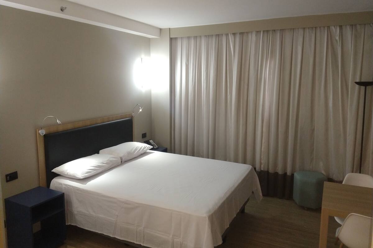 Une chambre d’hôtel comprenant un lit, une table de chevet, une lampe, des rideaux et un bureau avec une chaise.
