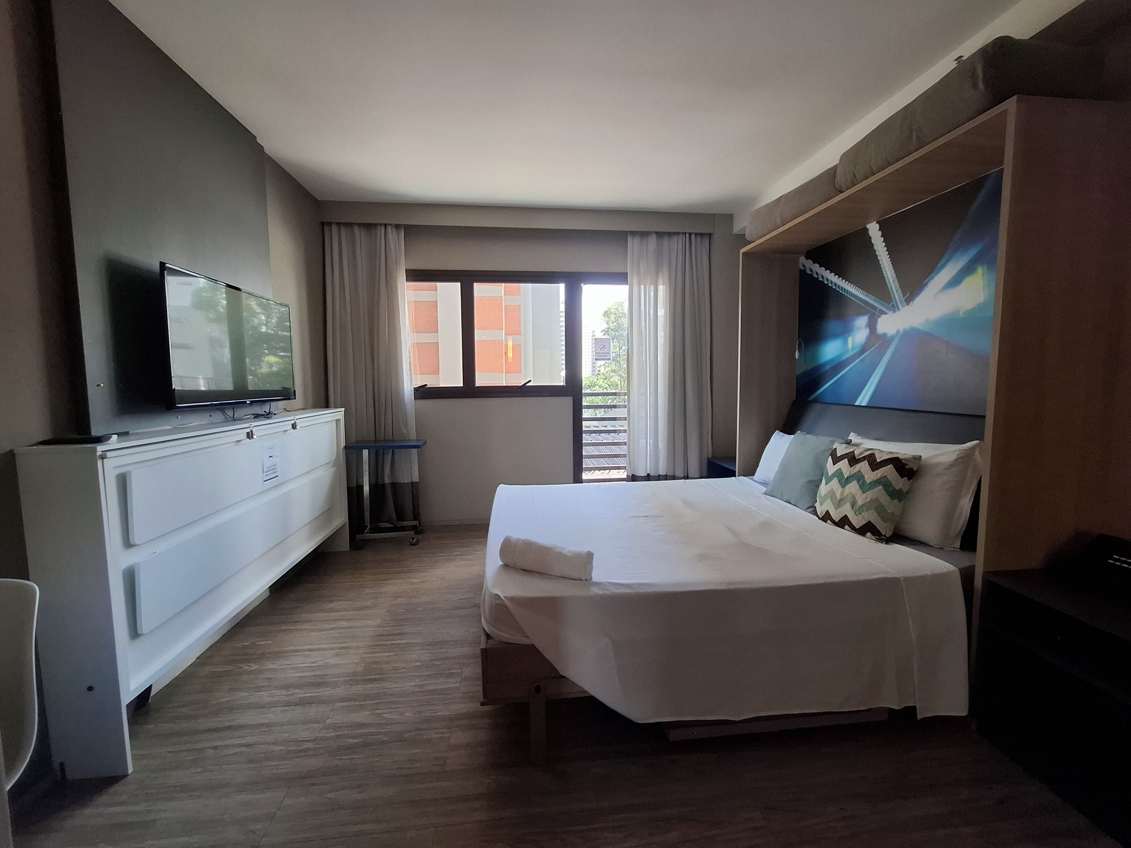 Habitación de hotel con cama, televisión, cómoda, ventana con cortinas y vistas al exterior.