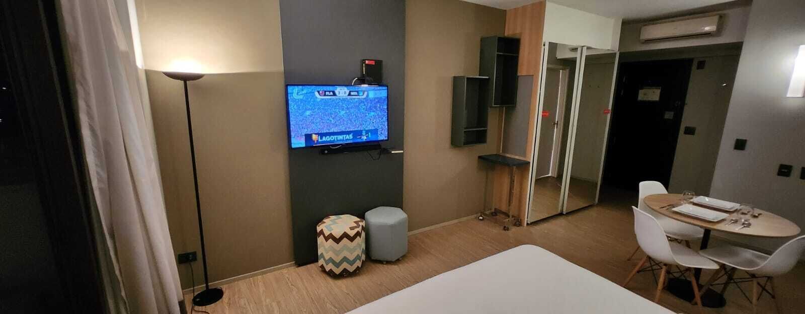 A hotel room with a television, a small ottoman, a dining table with chairs, and a wardrobe.