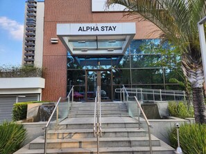 The entrance to Alpha Stay, featuring a modern glass facade, a set of stairs with metal railings, and a brick building facade. - Alpha Stay (Barueri)