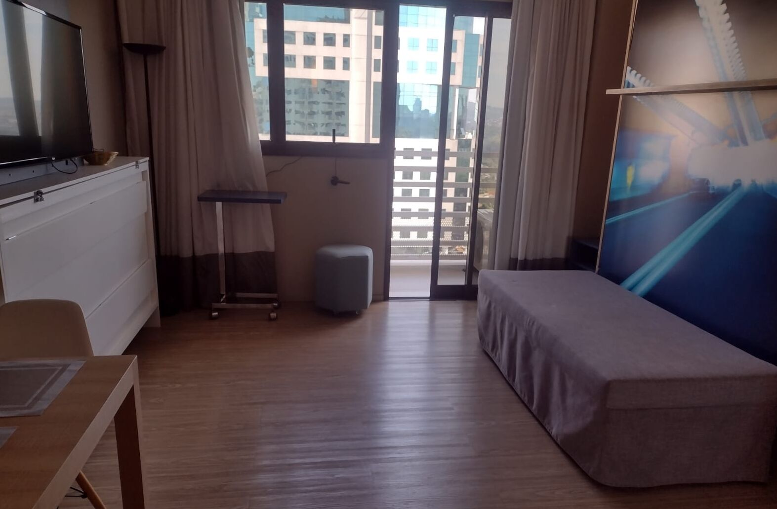 A hotel room with a bed, a desk, a television, a window with a view of buildings, and a curtain.