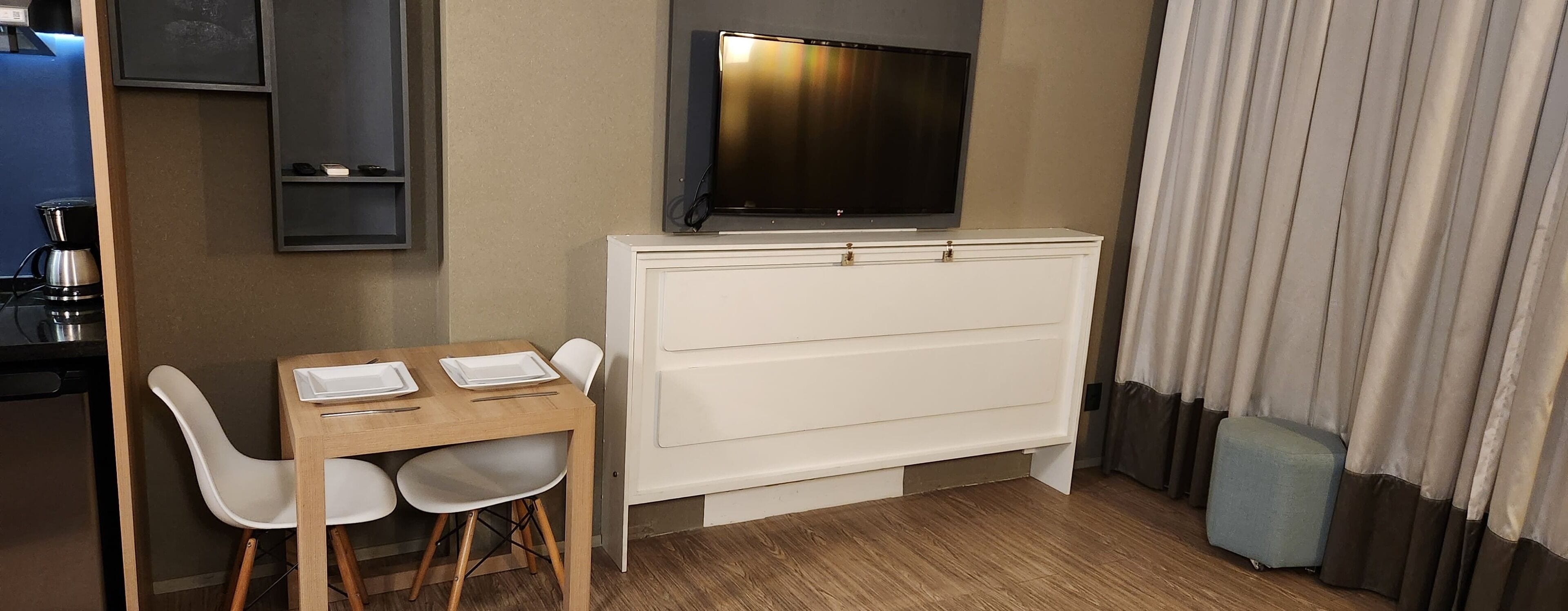 A small, modern room with a wooden dining table, two white chairs, a flat-screen TV mounted on a white cabinet, and a window with curtains.