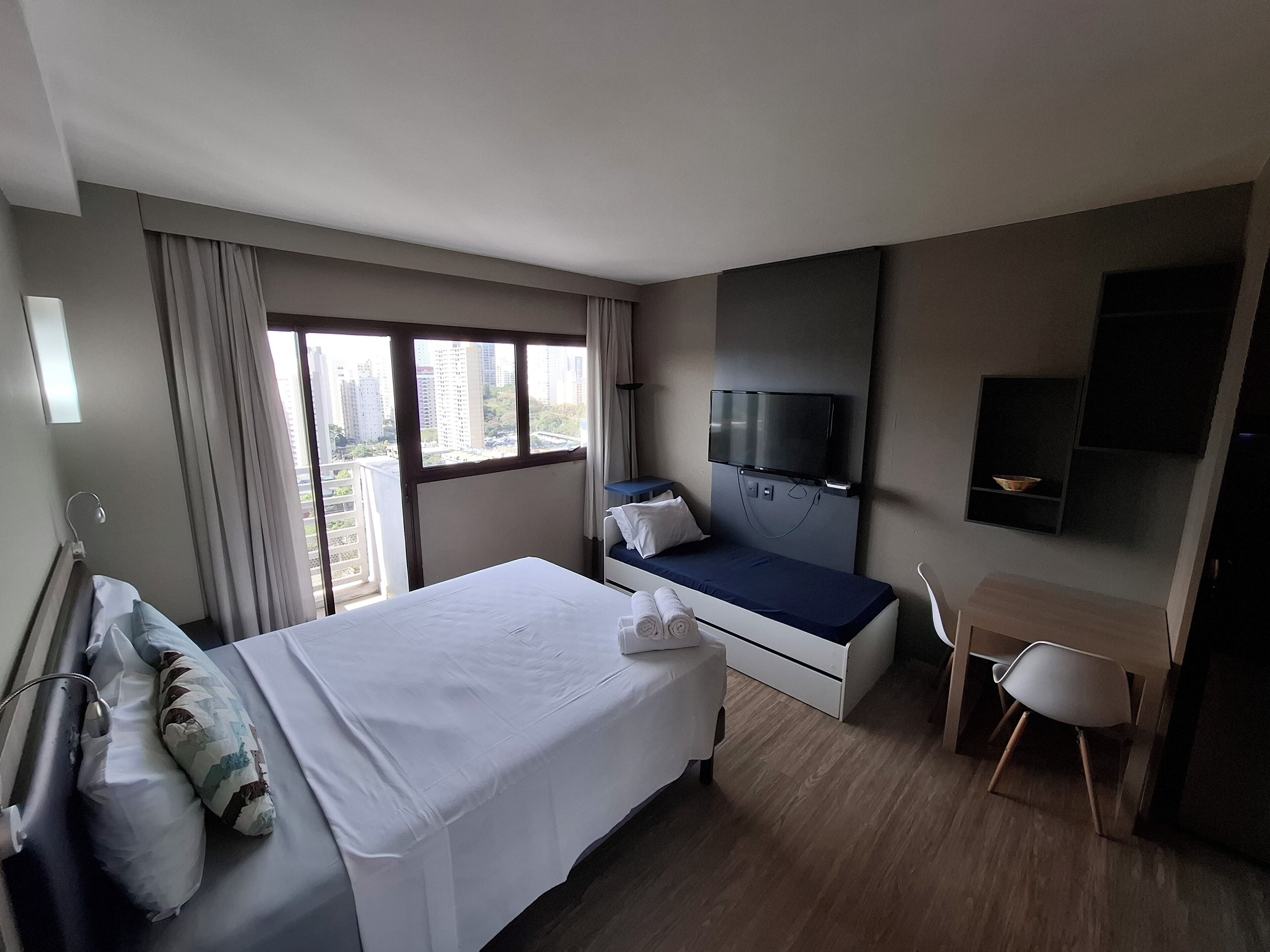 Una habitación de hotel con cama, mesita de noche, televisor, escritorio, silla y ventanas amplias.