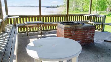 Een overdekt terras met een tafel, bankjes, een bakstenen barbecue en uitzicht op een waterpartij.
