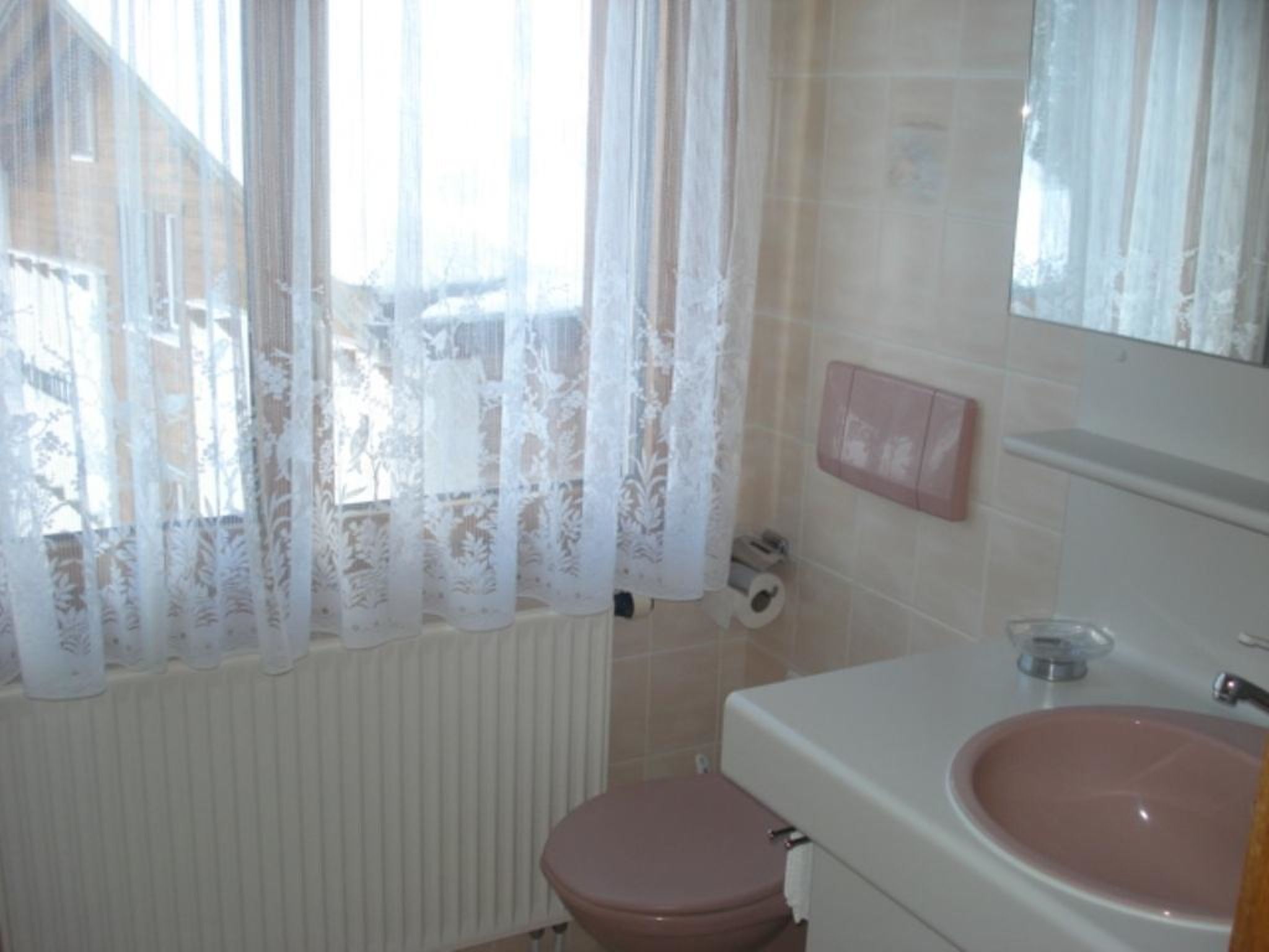 Un bagno con il water, un lavandino, uno specchio e una finestra con tenda in pizzo.
