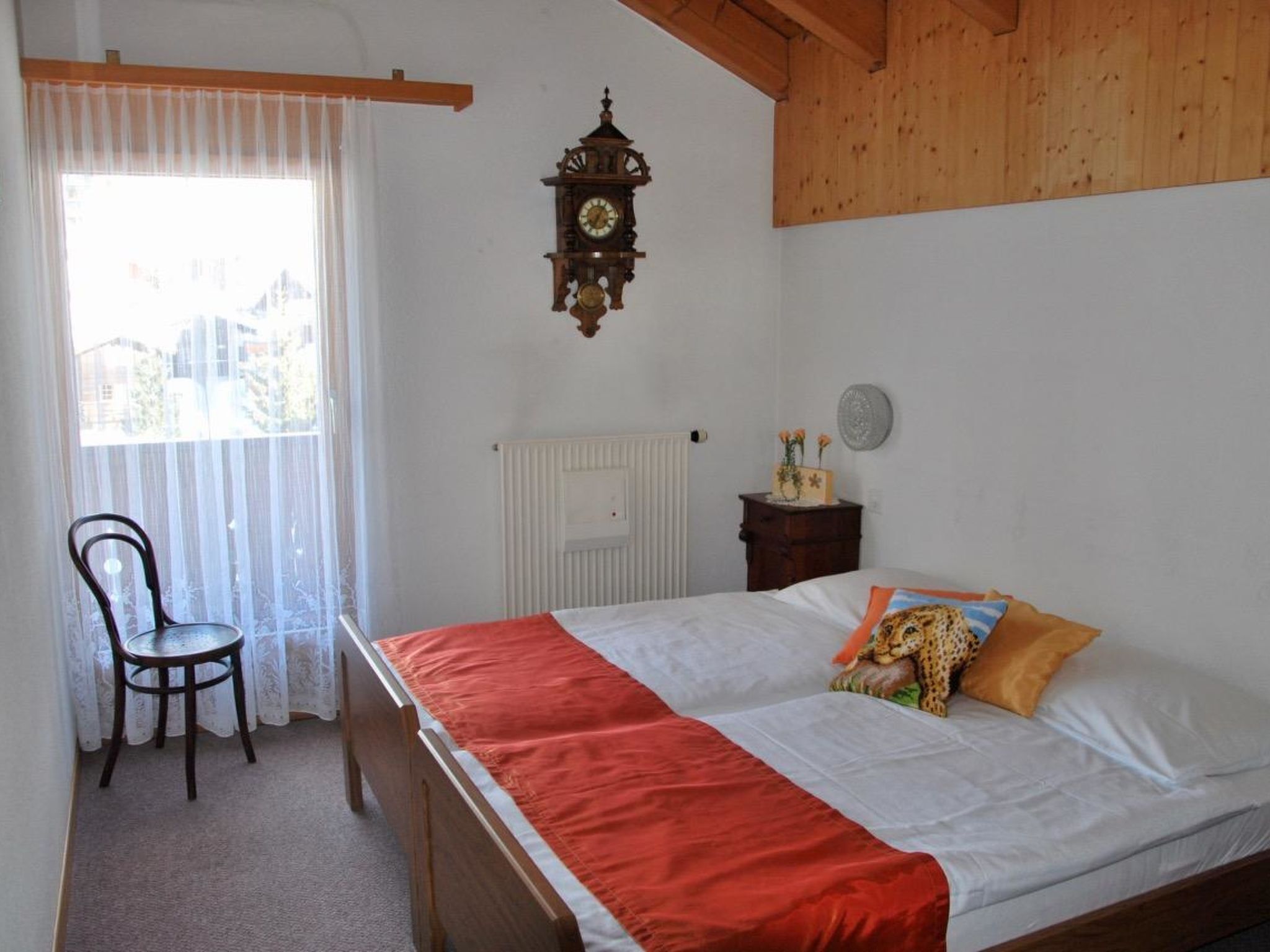 A bedroom with a wooden bed, a red bedspread, a chair, a wall clock, a radiator, and a window with curtains.