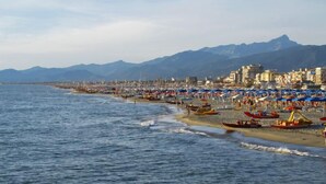 Una playa costera con numerosos sombrillas y barcos, un cielo despejado y montañas al fondo.