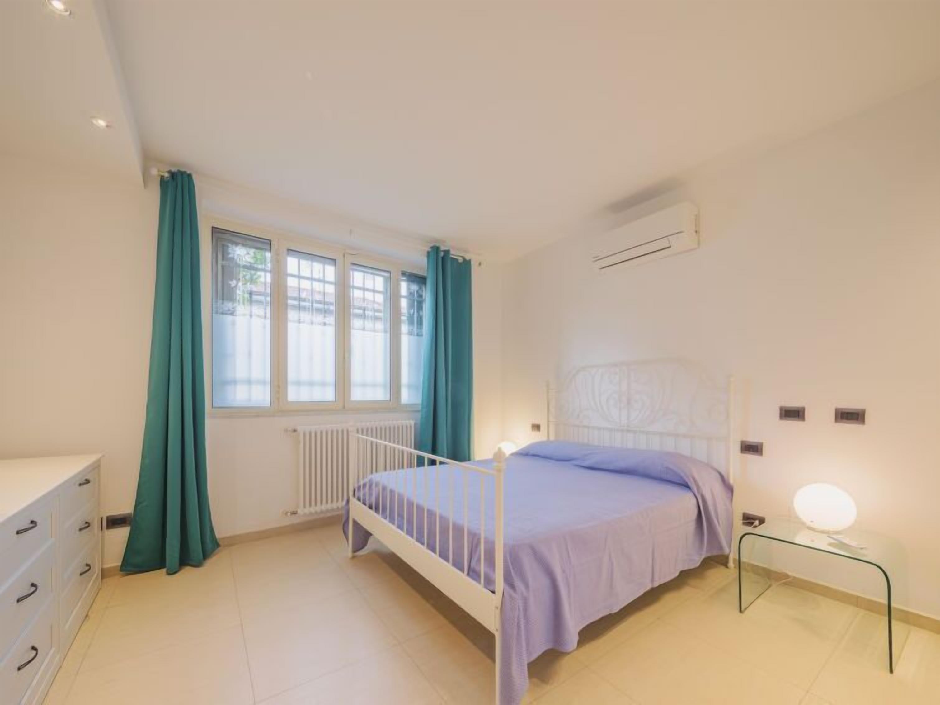 A bedroom with a bed, a dresser, a radiator, a window with curtains, and a bedside table with a lamp.