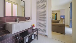 Un baño moderno con un espejo grande, un lavabo blanco y baldosas oscuras. Se ve un perchero y una pequeña mesa con un florero.