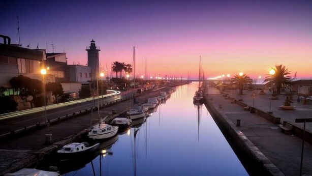 Una marina al atardecer con barcos amarrados a lo largo del agua, un faro y una hilera de edificios con luces encendidas.