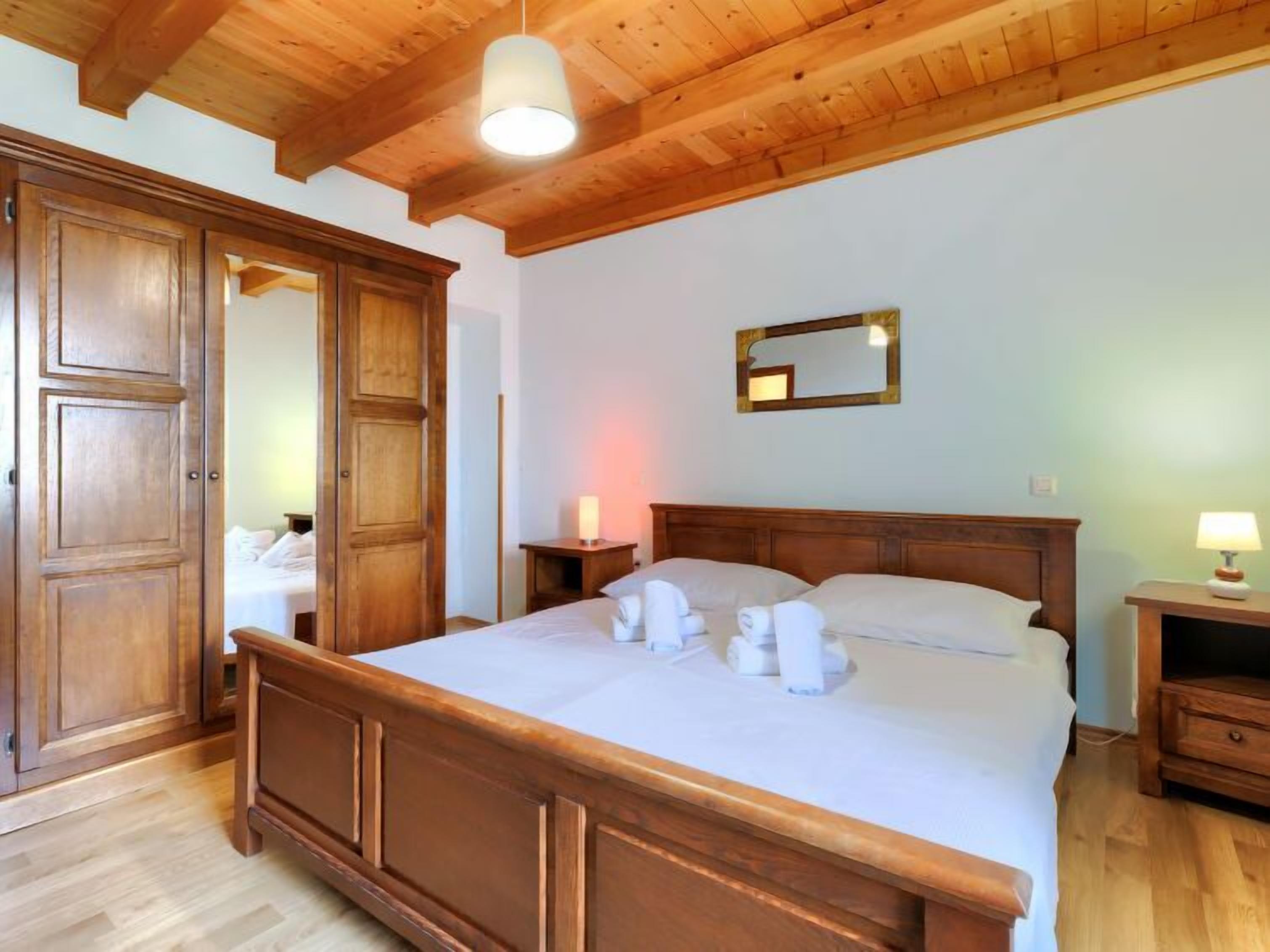 A wooden-framed bed with white linens and towels, a wooden wardrobe, a bedside table with a lamp, and a mirror on the wall.