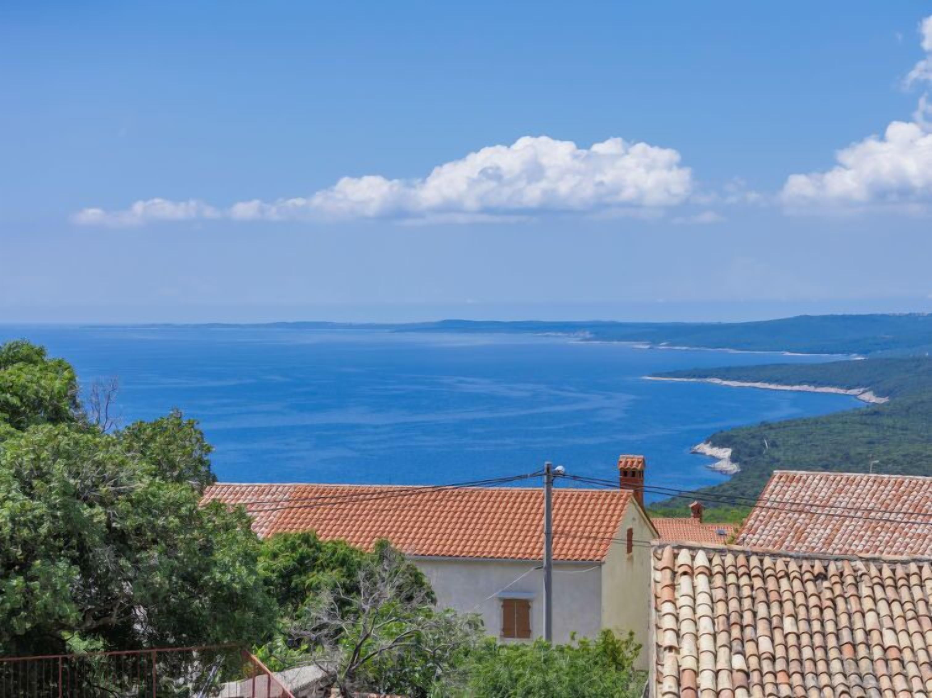 A coastal view with terracotta-roofed buildings, a clear blue sea, and distant landforms.