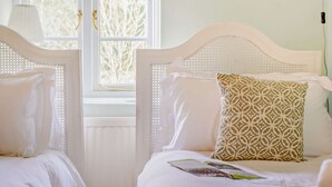A room with two beds, each with a white headboard and a patterned pillow. There is a window with a view of trees outside. - Characterful holiday cottage in the Cotswolds - Wren Cottage (Stow-on-the-Wold)