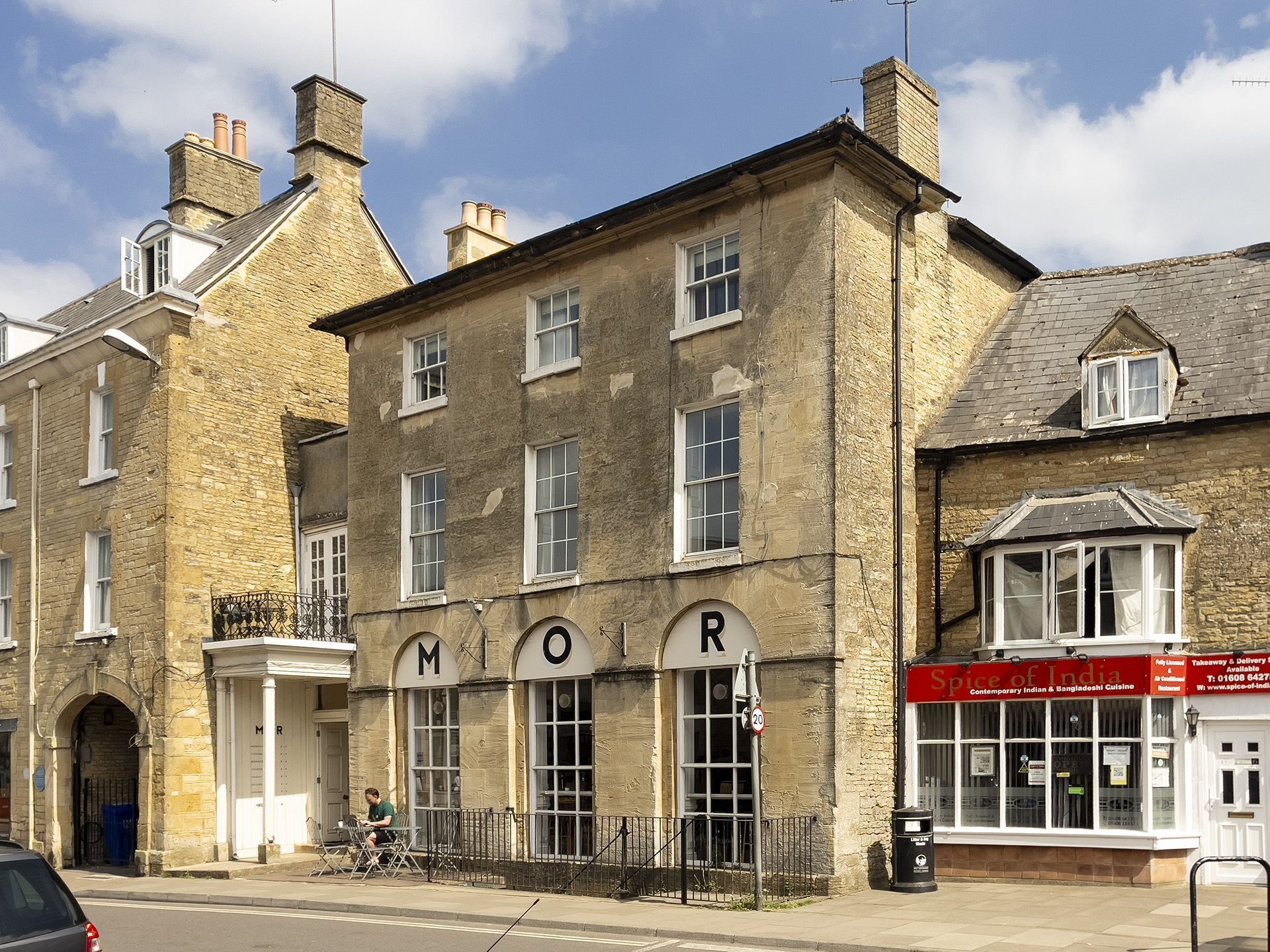 A historic building with a prominent 'MOR' sign, a restaurant named 'Taste of India', and a street view with a parked car and a person sitting outside.