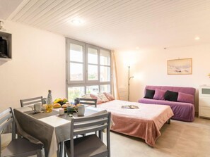 Ein gemütliches Zimmer mit Esstisch, Stühlen, einem Bett mit rosa Bettdecke, einem violetten Sofa und einem großen Fenster.
