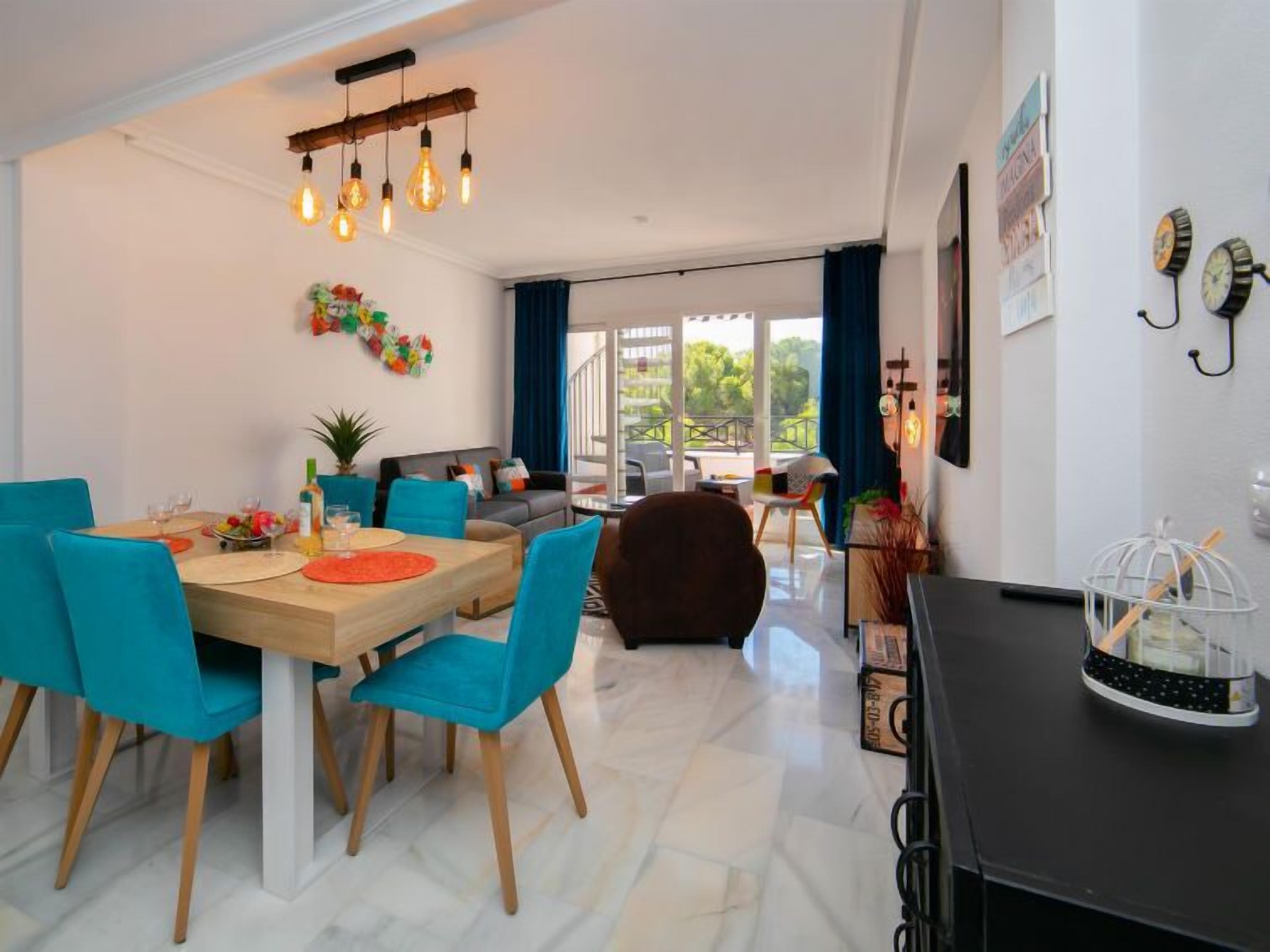 Een moderne woonkamer met een eethoek, met een houten tafel, blauwe stoelen en een hanglamp.