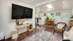 Een ruime woonkamer met een grote flatscreen televisie, houten vloeren, een eethoek met een ronde tafel, en een keuken op de achtergrond.