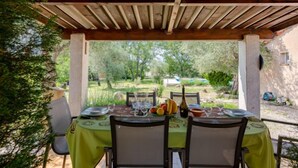 An outdoor dining area with a wooden pergola, a table set for two with chairs, and a view of a garden and trees in the background.
