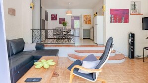 A modern living room with a sofa, a wooden table with a flower vase, a chair, and a dining area in the background.
