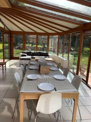 Enjoy meals in this dining area with a long table and comfortable seating, surrounded by a glass-enclosed patio with a garden view.