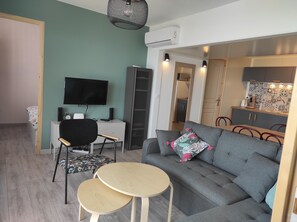 Un salon moderne avec un canapé gris, une petite table ronde, un téléviseur et, au fond, un coin cuisine.