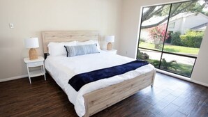 Un dormitorio con una cama grande, dos mesitas de noche con lámparas y vistas a un jardín a través de un ventanal.