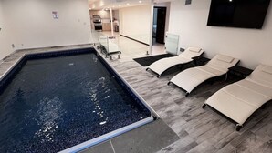 Ein moderner Poolbereich im Innenbereich mit Liegestühlen, einer Küche im Hintergrund und einem flachbildschirmigen Fernseher an der Wand.