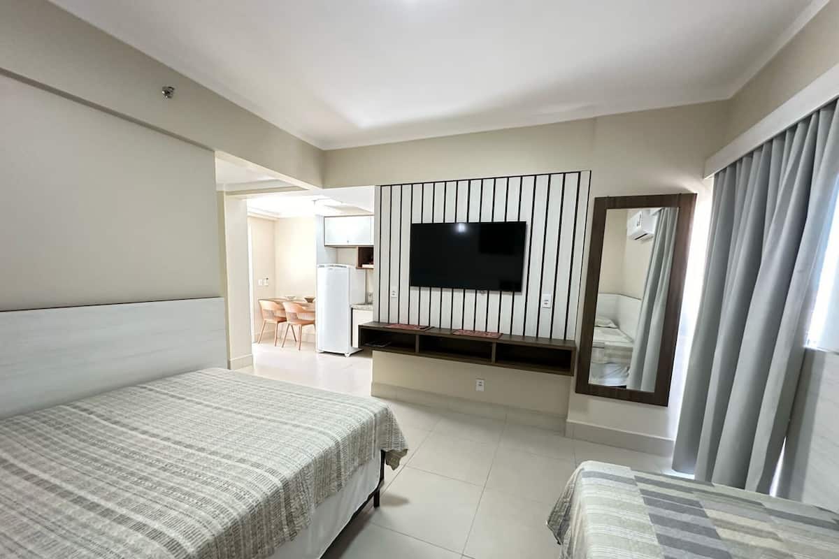 A hotel room with a large bed, a smaller bed, a TV mounted on the wall, a mirror, and a kitchenette in the background.