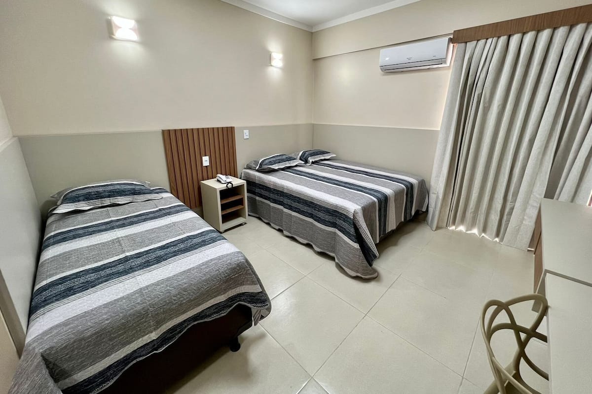 A hotel room with two beds, a small table, a nightstand, a wall-mounted air conditioner, and a window with curtains.
