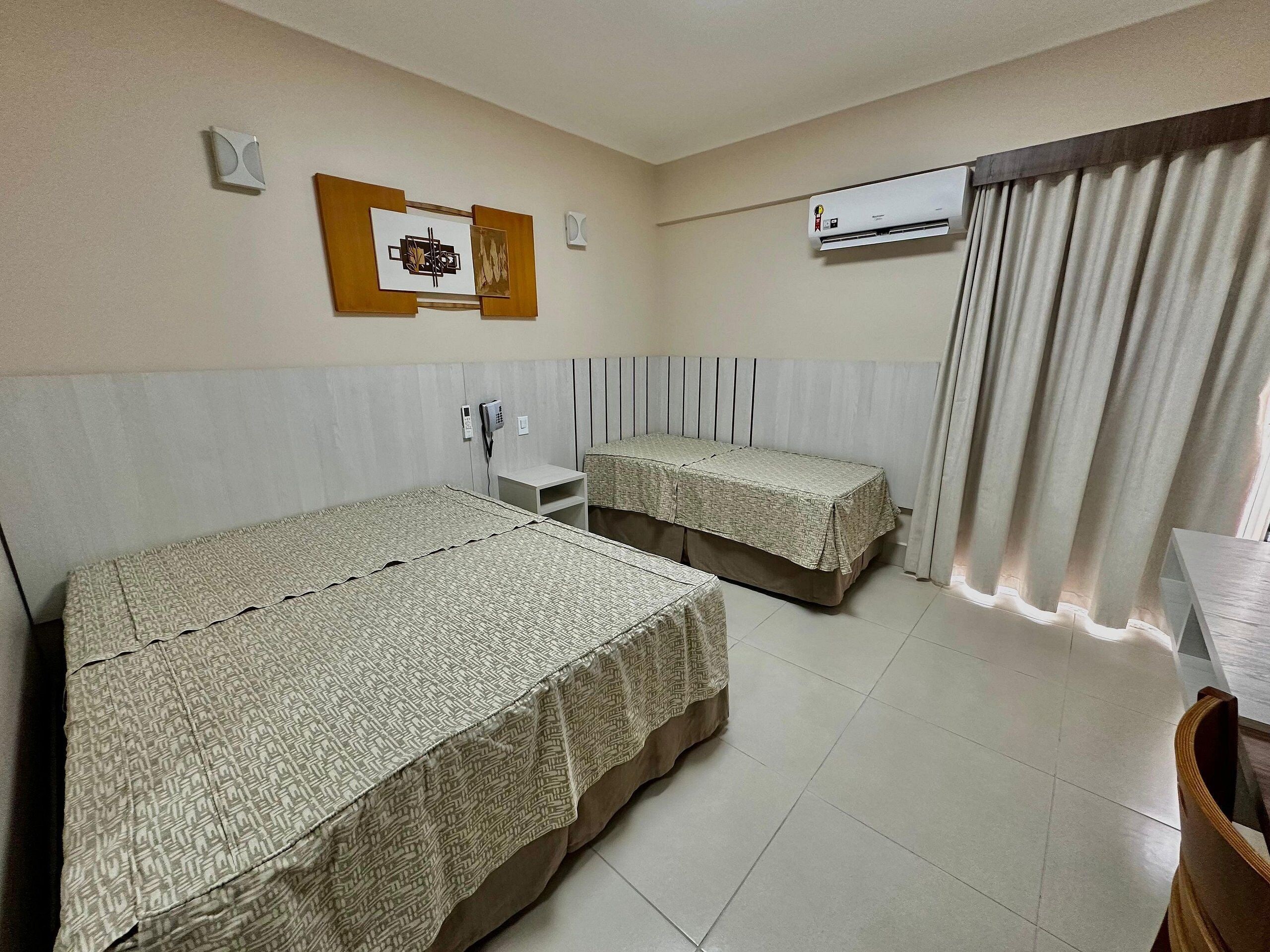A hotel room with two beds, a bedside table, a telephone, an air conditioning unit, and a window with curtains.