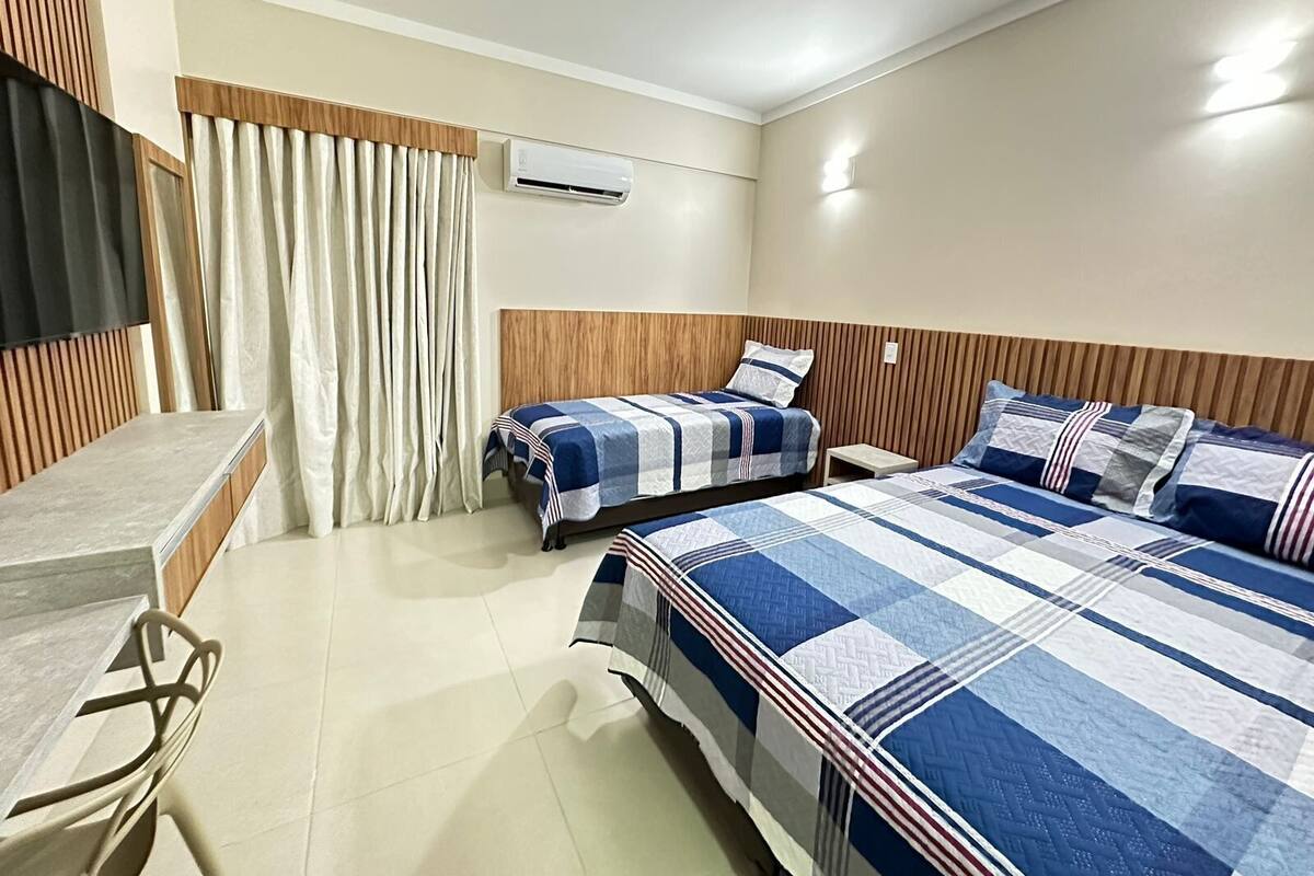A hotel room with two beds, a desk, a chair, an air conditioning unit, and a window with curtains.