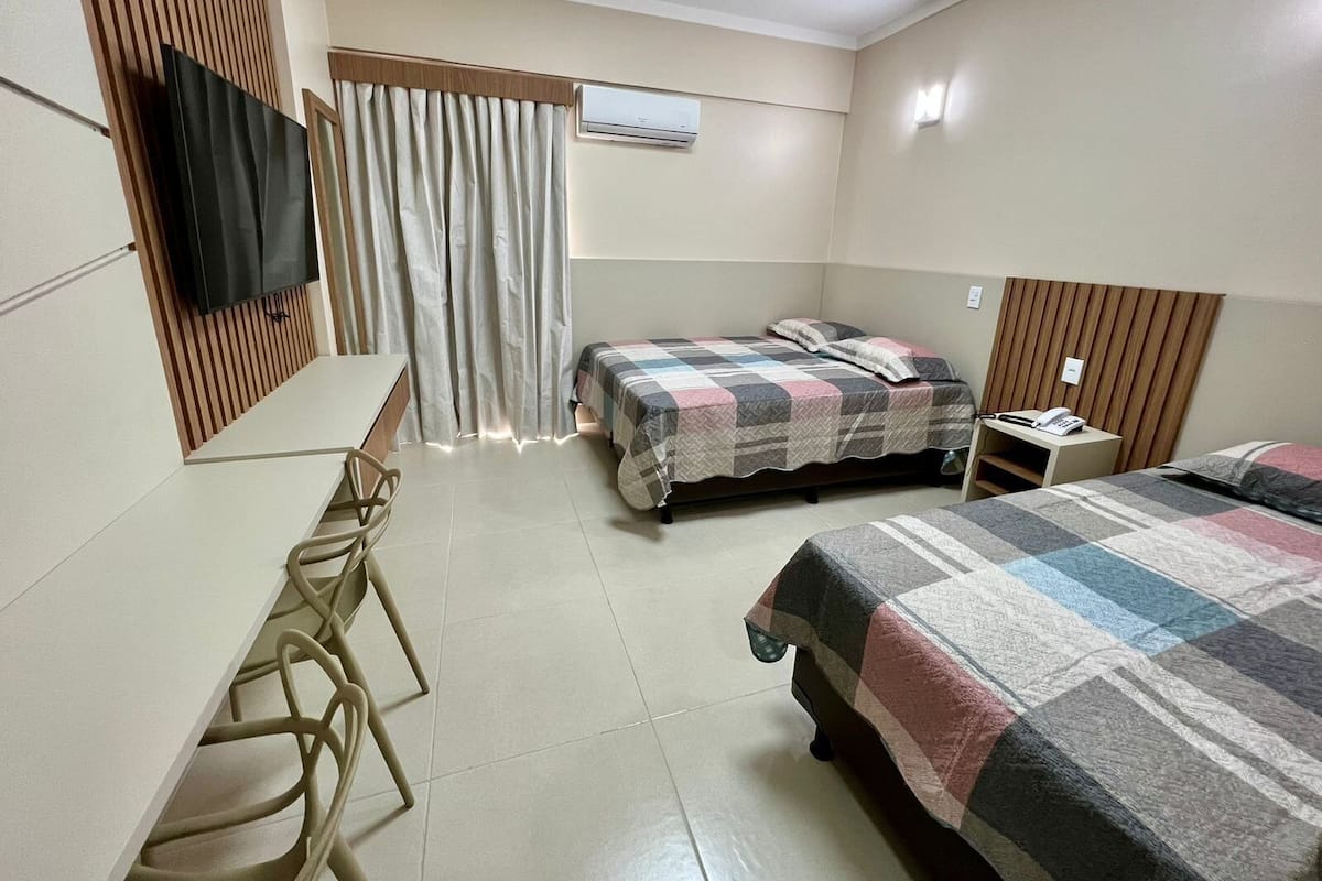 A hotel room with a bed, a desk with a chair, a TV, and an air conditioning unit.