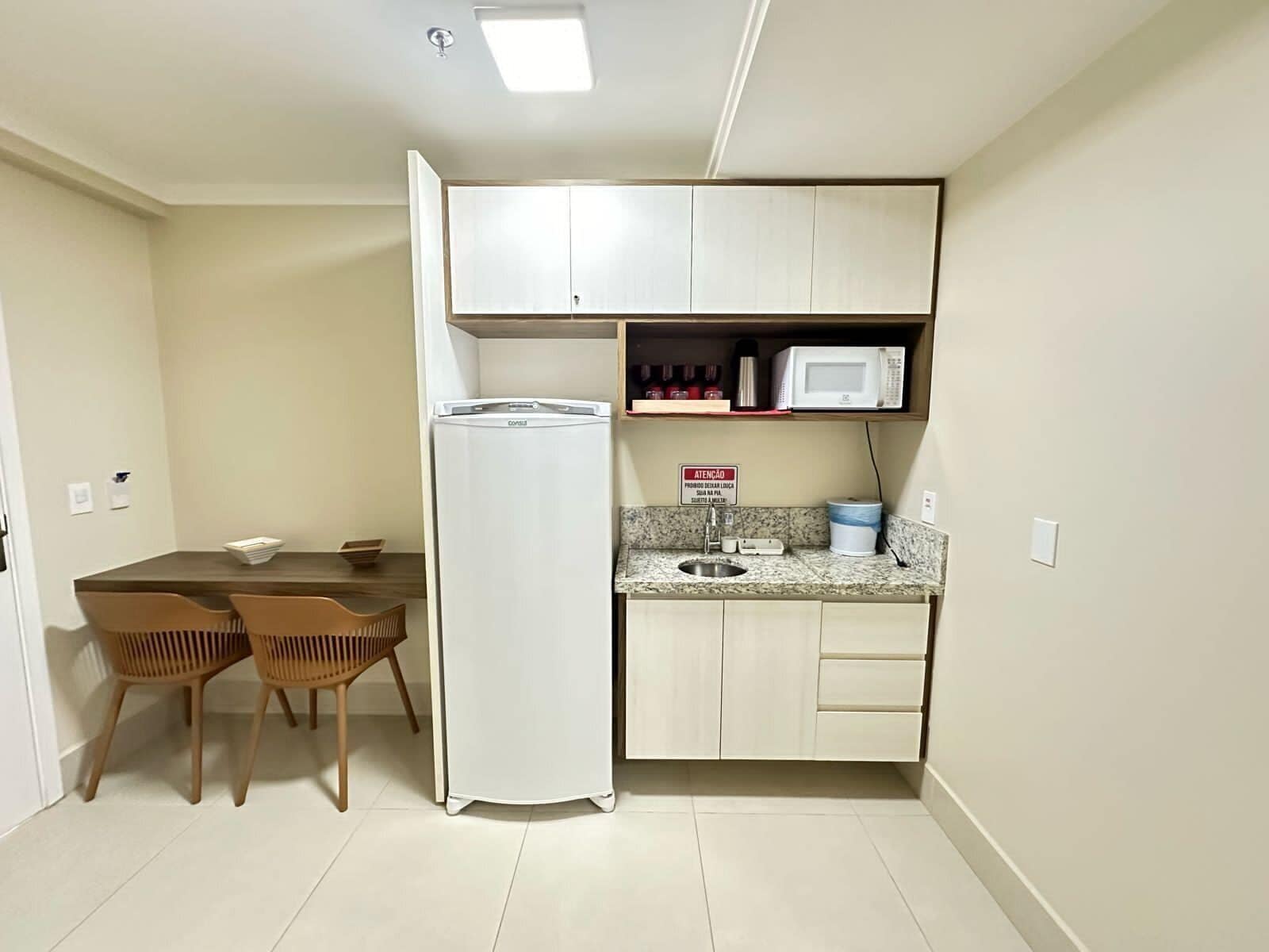 Cocina compacta con refrigerador, microondas y lavaplatos. Hay una mesa de comedor con sillas y una pequeña estantería con objetos.