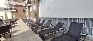 A sunlit outdoor seating area with lounge chairs and tables, a building with a brick facade, and a white wall with a window. - Di Roma Spazzio Via Caldas (Caldas Novas)