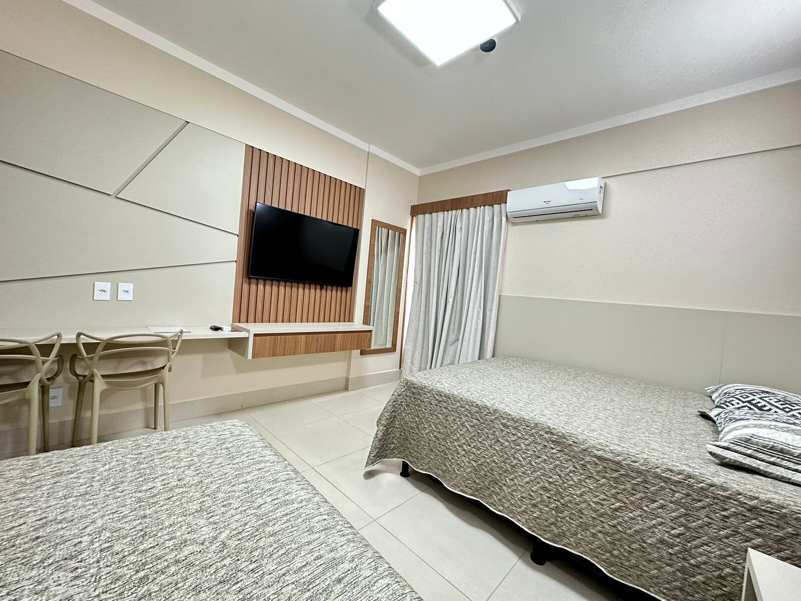 A hotel room with a bed, a TV mounted on the wall, a desk with chairs, and an air conditioning unit.