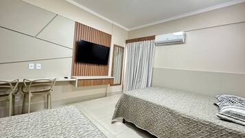 A hotel room with a bed, a TV mounted on the wall, a desk with chairs, and an air conditioning unit.