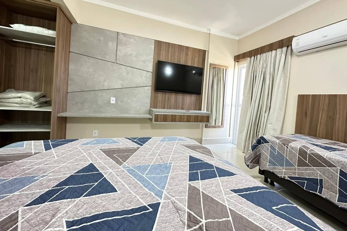 A hotel room with a large bed, a TV mounted on the wall, a wardrobe, and an air conditioning unit.