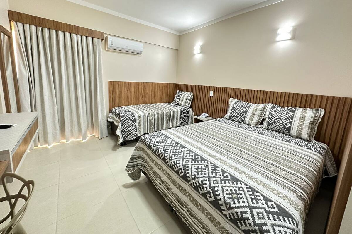 A hotel room with two beds, a desk, a chair, a window with curtains, an air conditioning unit, and a wall-mounted light.