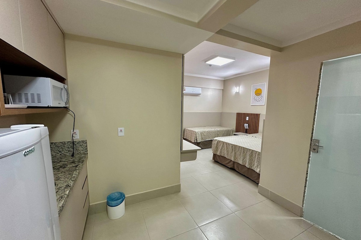 A hotel room with a kitchenette, a bed, a bedside table, a trash bin, and a door.