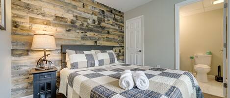 A bedroom with a wooden wall, a bed with a quilt, a nightstand, and a door leading to a bathroom.