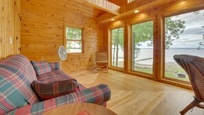 A wooden cabin interior with a plaid sofa, a fan, and large windows overlooking a body of water. - Lake Champlain Vacation Rental with Boat Dock! (St. Albans Town)