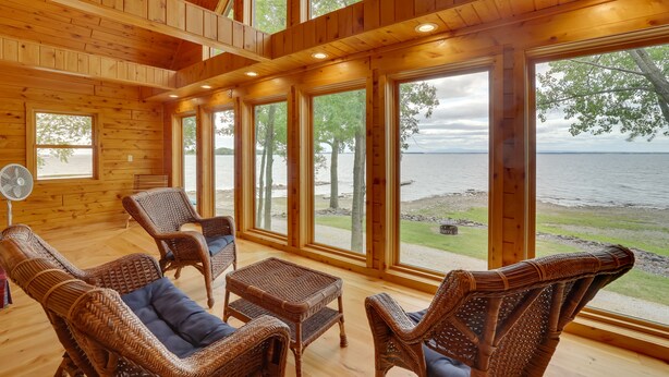 A wooden cabin with large windows overlooking a lake, wicker furniture, and a fan. - Lake Champlain Vacation Rental with Boat Dock! (St. Albans Town)