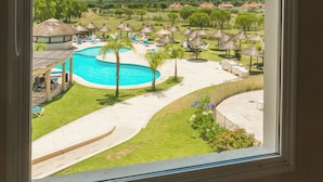 On aperçoit une piscine, des chaises longues et des parasols depuis une fenêtre.