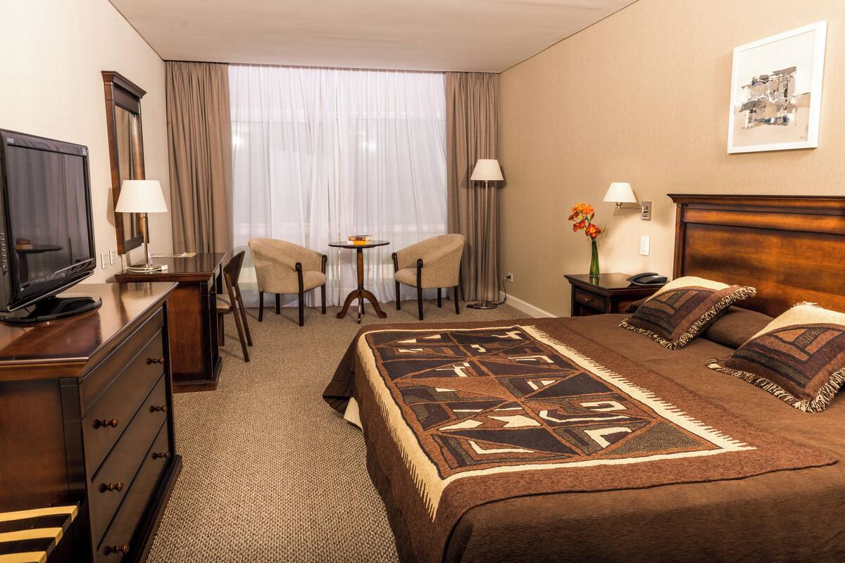 Ein Hotelzimmer mit Bett, Fernseher, Schreibtisch, zwei Stühlen, einem kleinen Tisch und einem Fenster mit Vorhängen.