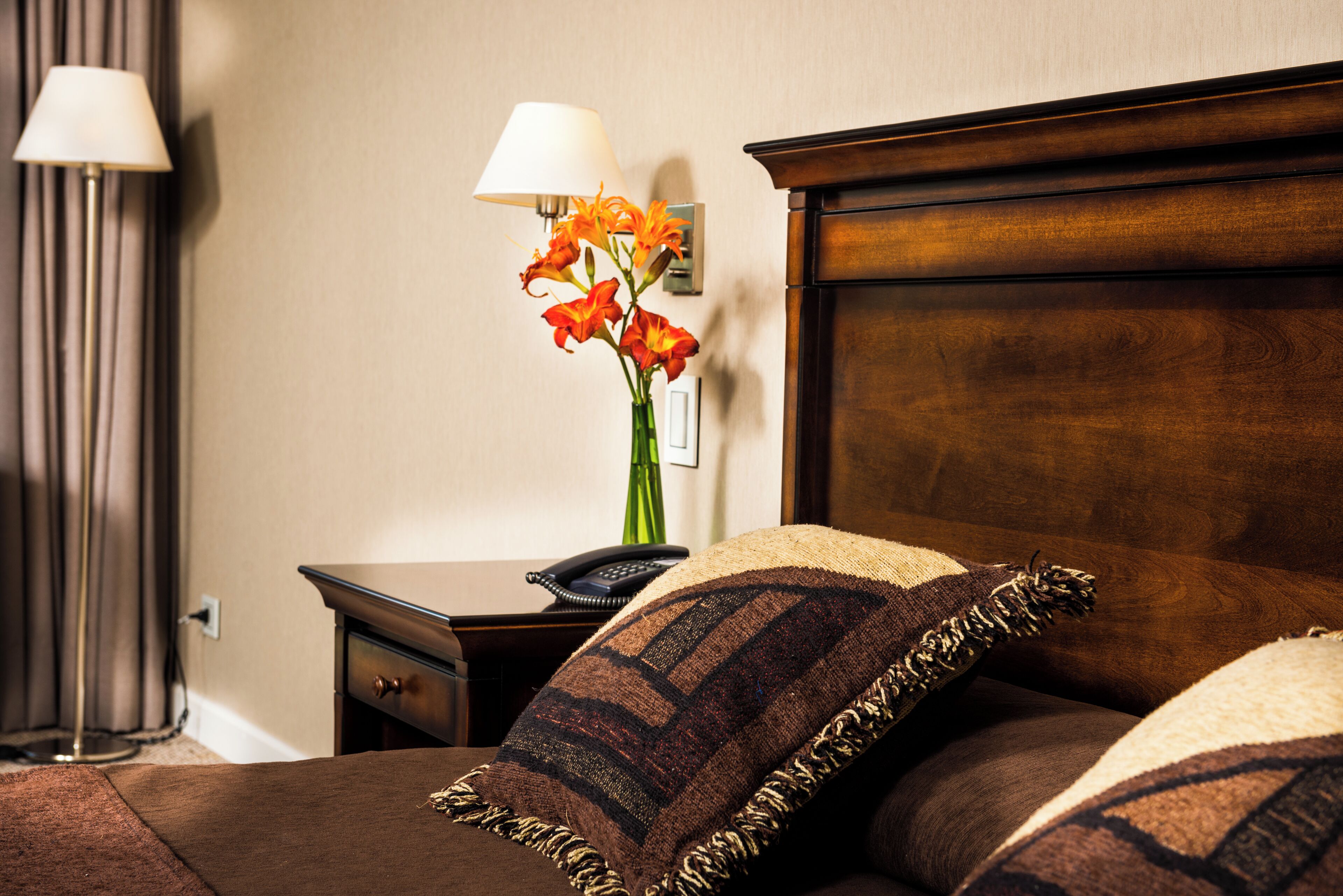 A neatly made bed with a brown headboard, a bedside table with a lamp and a vase of flowers, and a wall-mounted light fixture.