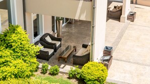 Un espace extérieur moderne aménagé avec des meubles en osier et une table en verre, entouré de verdure et bordé d’un bâtiment doté de grandes fenêtres.