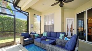 Un espacio de patio cubierto con mobiliario de mimbre, un tapete azul a rayas y ventanas amplias que ofrecen vistas a la vegetación.