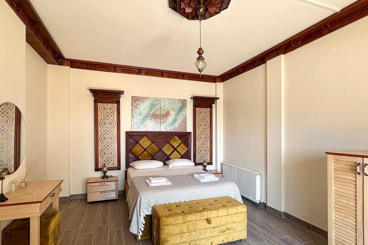 A bedroom with a wooden headboard, a bed with a patterned bedspread, a bedside table, a nightstand, a wooden bench, and a decorative ceiling light.